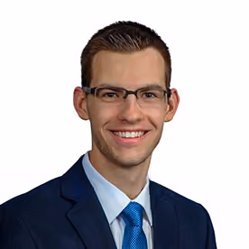 Smiling man with short hair and glasses wearing a dark suit, blue shirt, and blue tie.