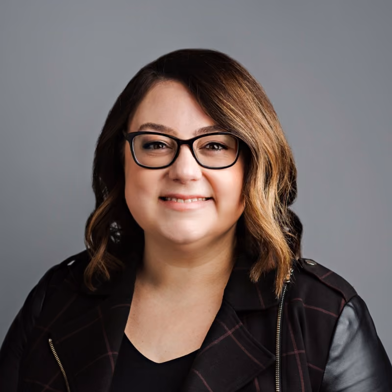 Smiling woman with shoulder-length brown hair, glasses, and a dark jacket against a gray background.