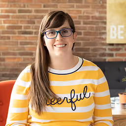 Smiling woman with long brown hair and glasses wearing a yellow and white striped sweater with cheerful text.