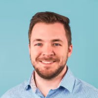 Smiling man with short brown hair and beard wearing a light blue collared shirt against teal background.