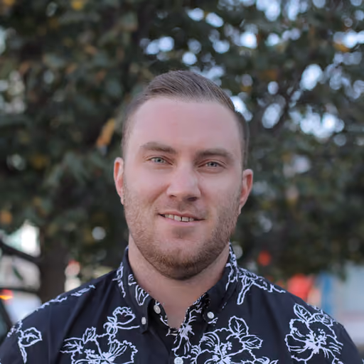 Man with short hair and beard wearing a black floral button-up shirt outdoors near trees.