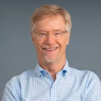 Smiling man with glasses and gray hair wearing a blue checkered shirt against a gray background.