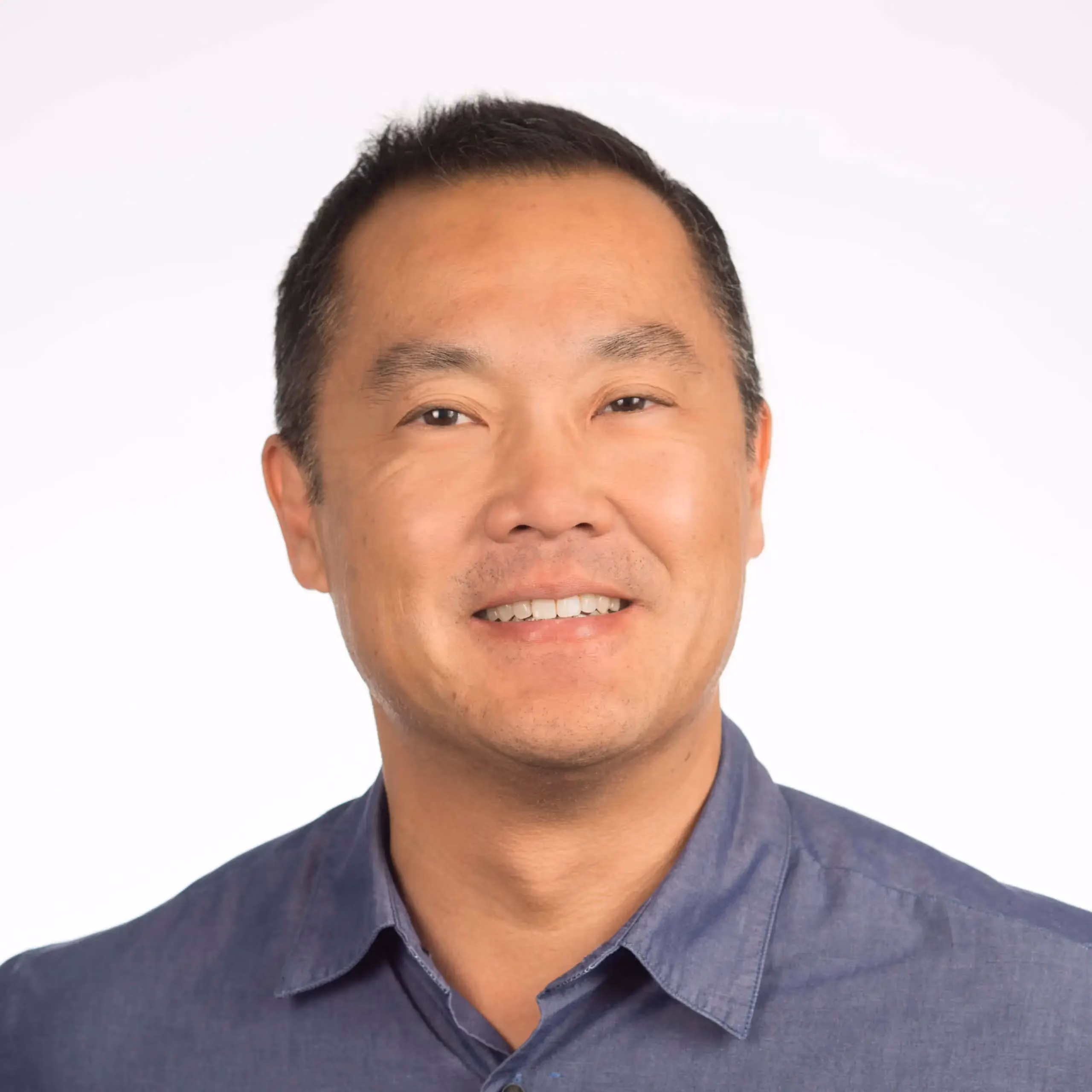 Smiling man with short black hair wearing a blue button-up shirt against a white background.