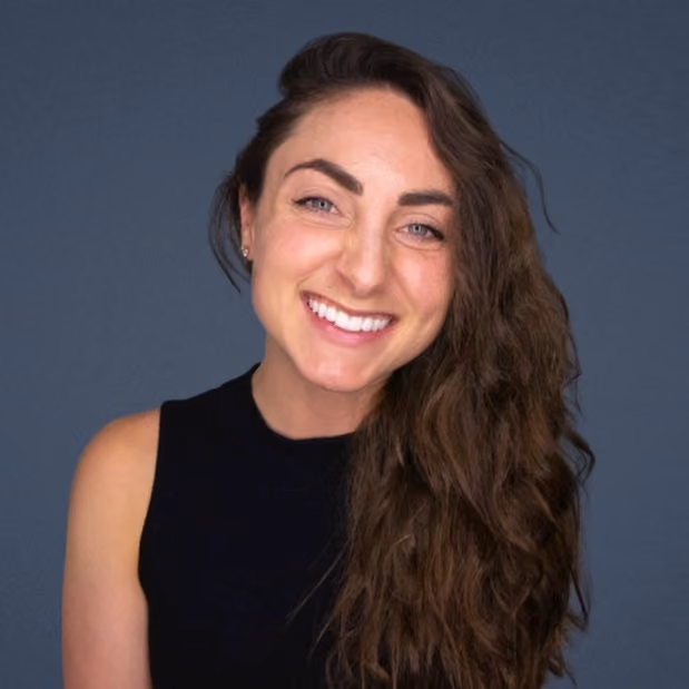 Smiling woman with long curly brown hair wearing a black sleeveless top against a gray background.