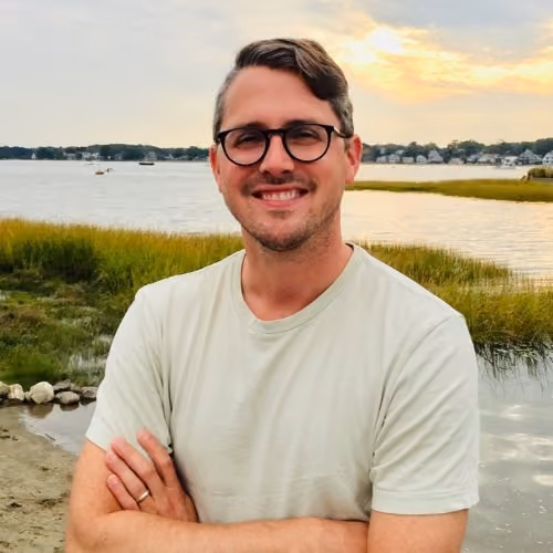 Man with glasses and light shirt smiling with arms crossed by a waterfront at sunset.