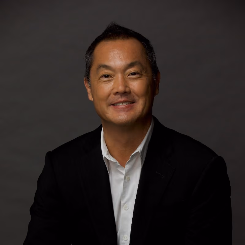 Smiling man in a black blazer and white shirt against a dark gray backdrop.