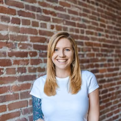 Smiling woman with long blonde hair, white shirt, and tattoo sleeve standing against a brick wall.