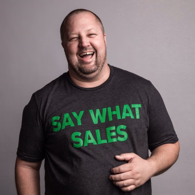 Smiling man wearing a dark shirt with green text saying Say What Sales on a gray background.