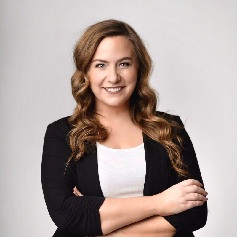 Smiling woman with wavy brown hair, wearing a white top and black blazer, arms crossed.