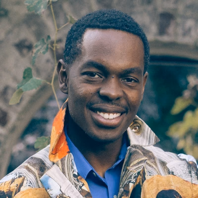 Smiling man with short hair wearing a patterned jacket and blue shirt outdoors near greenery.