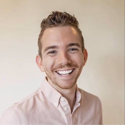 Smiling man with short curly hair and light beard wearing a light pink button-up shirt.
