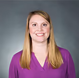 Smiling woman with long blonde hair wearing a purple V-neck blouse against a gray background.