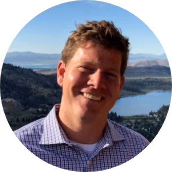 Smiling man in checkered shirt with mountains and lake in the background on a clear day.