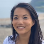 Smiling woman with long dark hair outdoors near water and rocky shore.