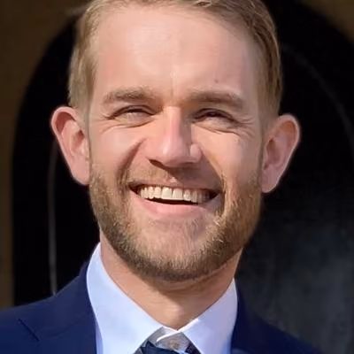 Smiling man with beard wearing a blue suit and tie against a dark background.