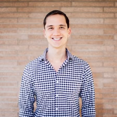 Smiling man wearing a black and white checkered shirt in front of a brick wall.