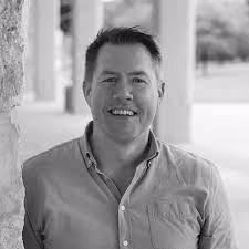 Smiling man with short dark hair wearing a buttoned shirt standing outdoors near columns.