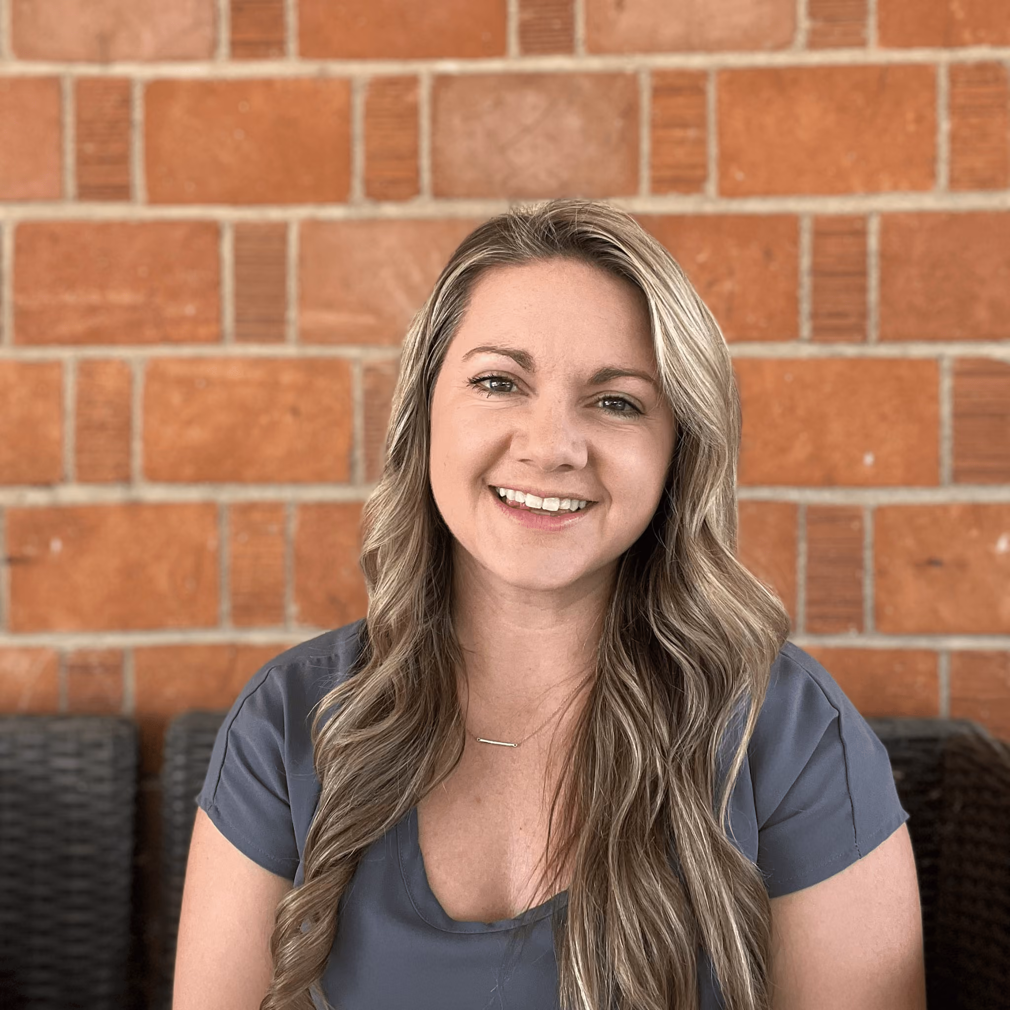 Smiling woman with long wavy blonde hair wearing a blue blouse seated against a brick wall.