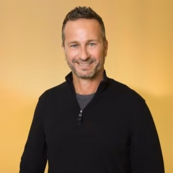 Smiling man with short hair and stubble wearing a black quarter-zip sweater on yellow background.