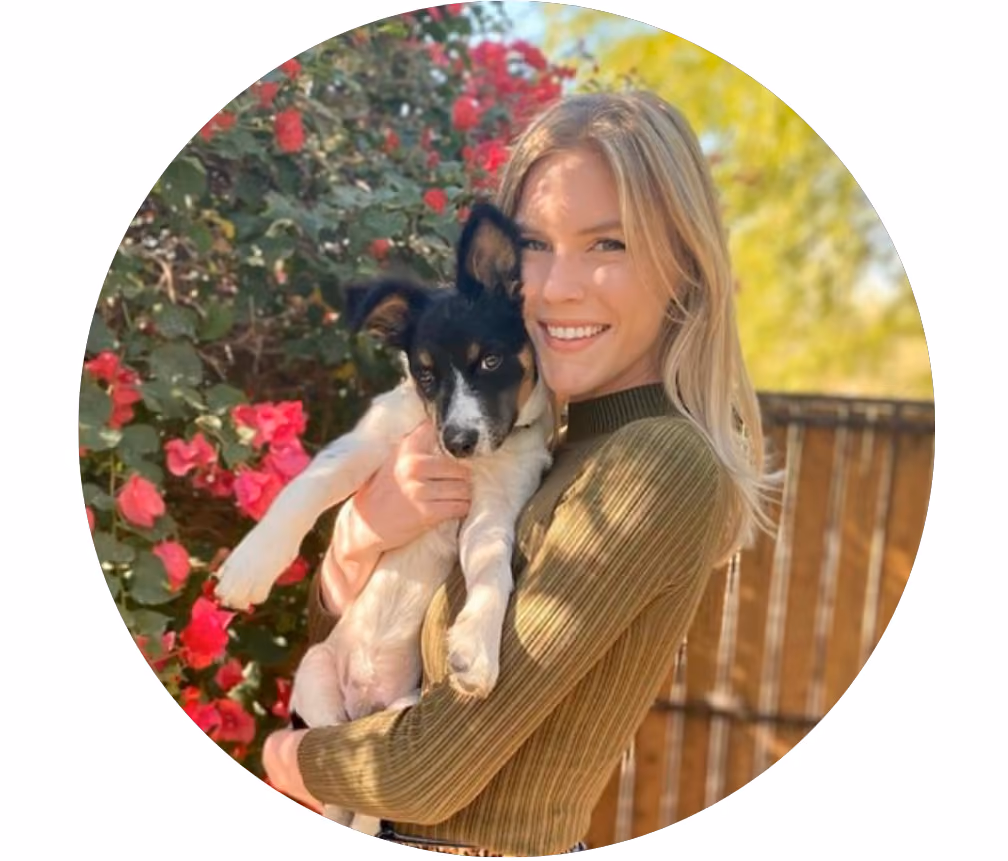 Smiling woman holding a black and white dog with red flowers and wooden fence behind.