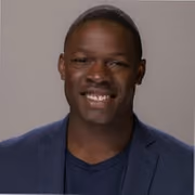 Smiling man wearing a dark blue blazer and navy shirt against a plain background.