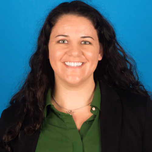 Smiling woman with long dark curly hair wearing a green shirt and black blazer on a blue background.