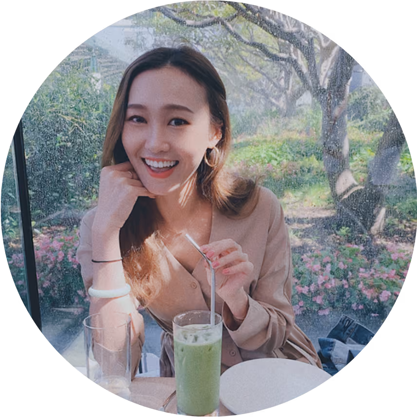 Smiling woman sitting at table with iced green drink holding a metal straw near a window with garden view.