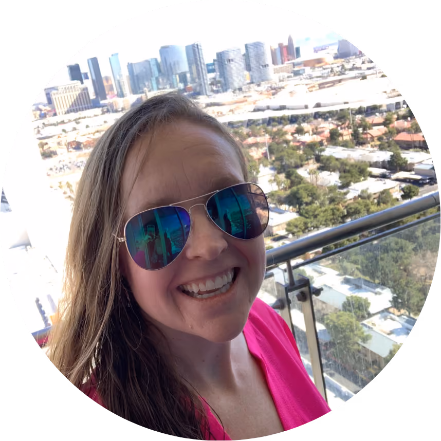 Woman wearing reflective sunglasses and a pink top smiling on a balcony with city skyline behind her.