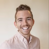 Smiling young man with short curly hair wearing a light pink button-up shirt against a neutral background.