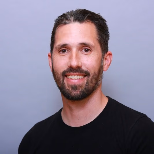 Smiling man with dark hair and beard wearing a black shirt against a light gray background.