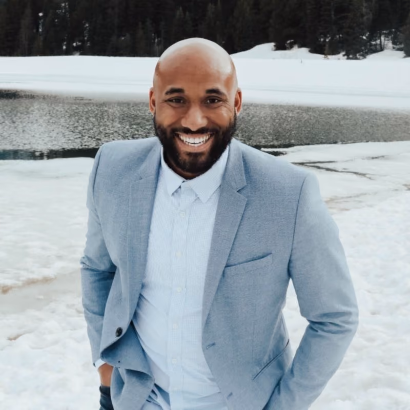 Smiling bald man with beard wearing light blue suit and shirt, standing outdoors on snow near water.