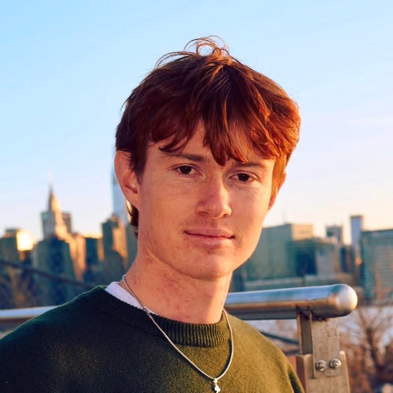 Young man with red hair and green sweater stands outdoors with city skyline at sunset in background.