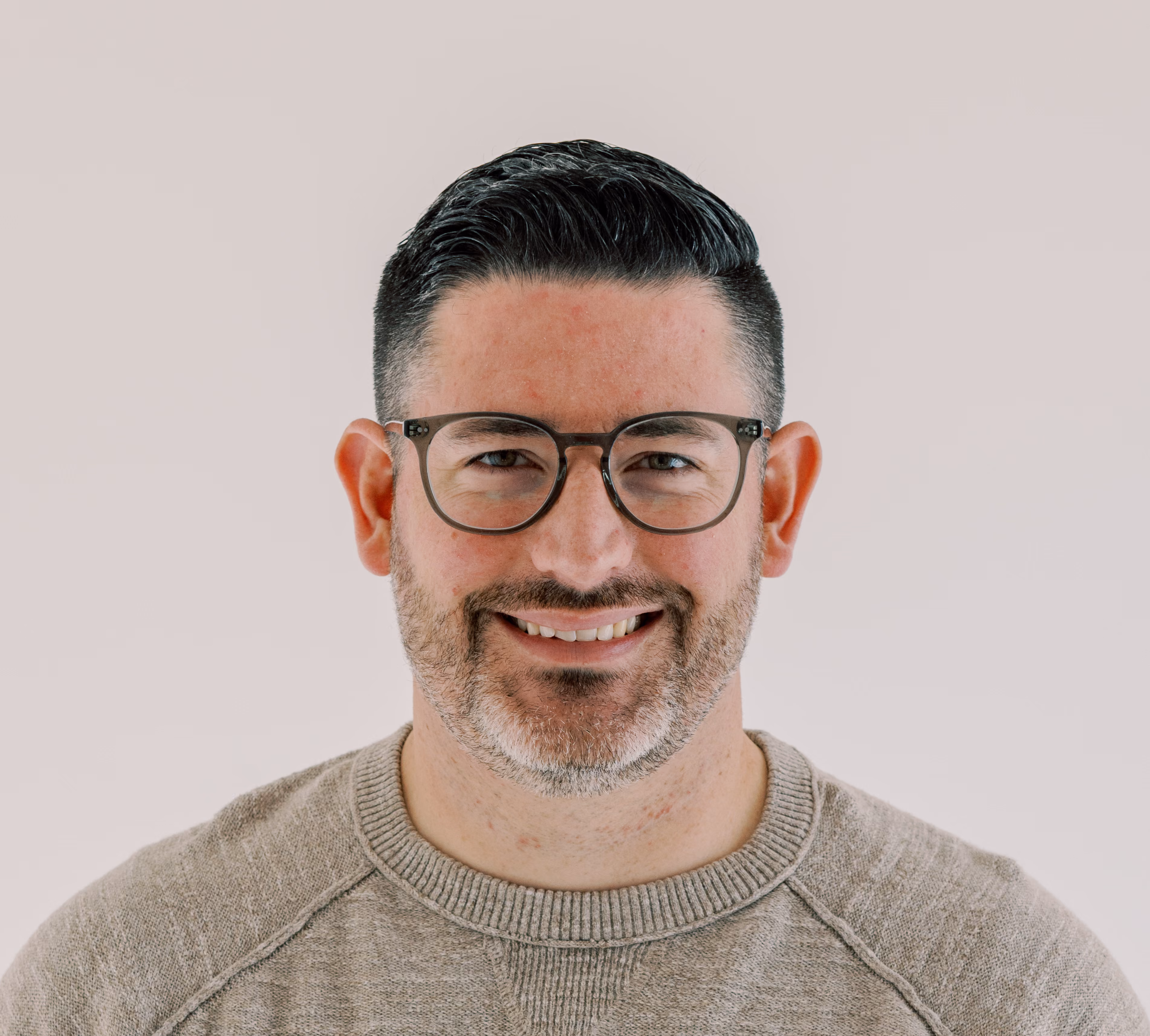 Smiling man with dark hair, glasses, and a beard wearing a light brown sweater against a plain background.