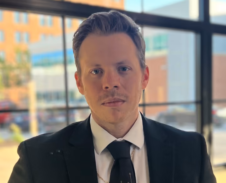 Man with light hair in a black suit and tie standing indoors with large windows and a cityscape behind him.