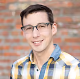 Young man with short dark hair, glasses, and a yellow plaid shirt smiling in front of a brick wall.