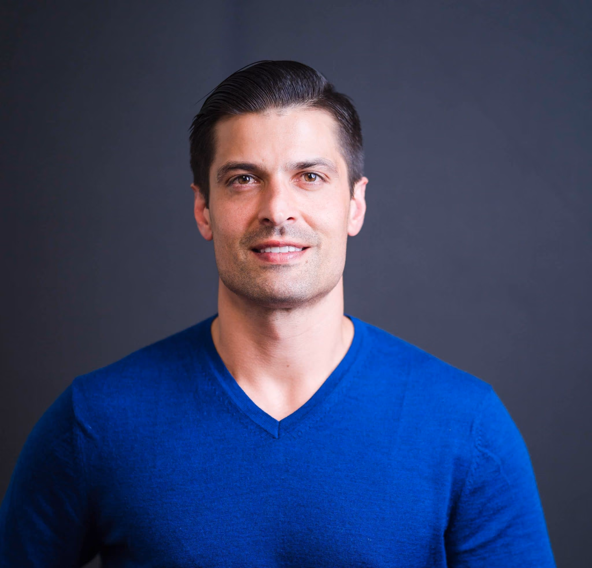 Smiling man with dark hair wearing a blue v-neck shirt against a dark background.