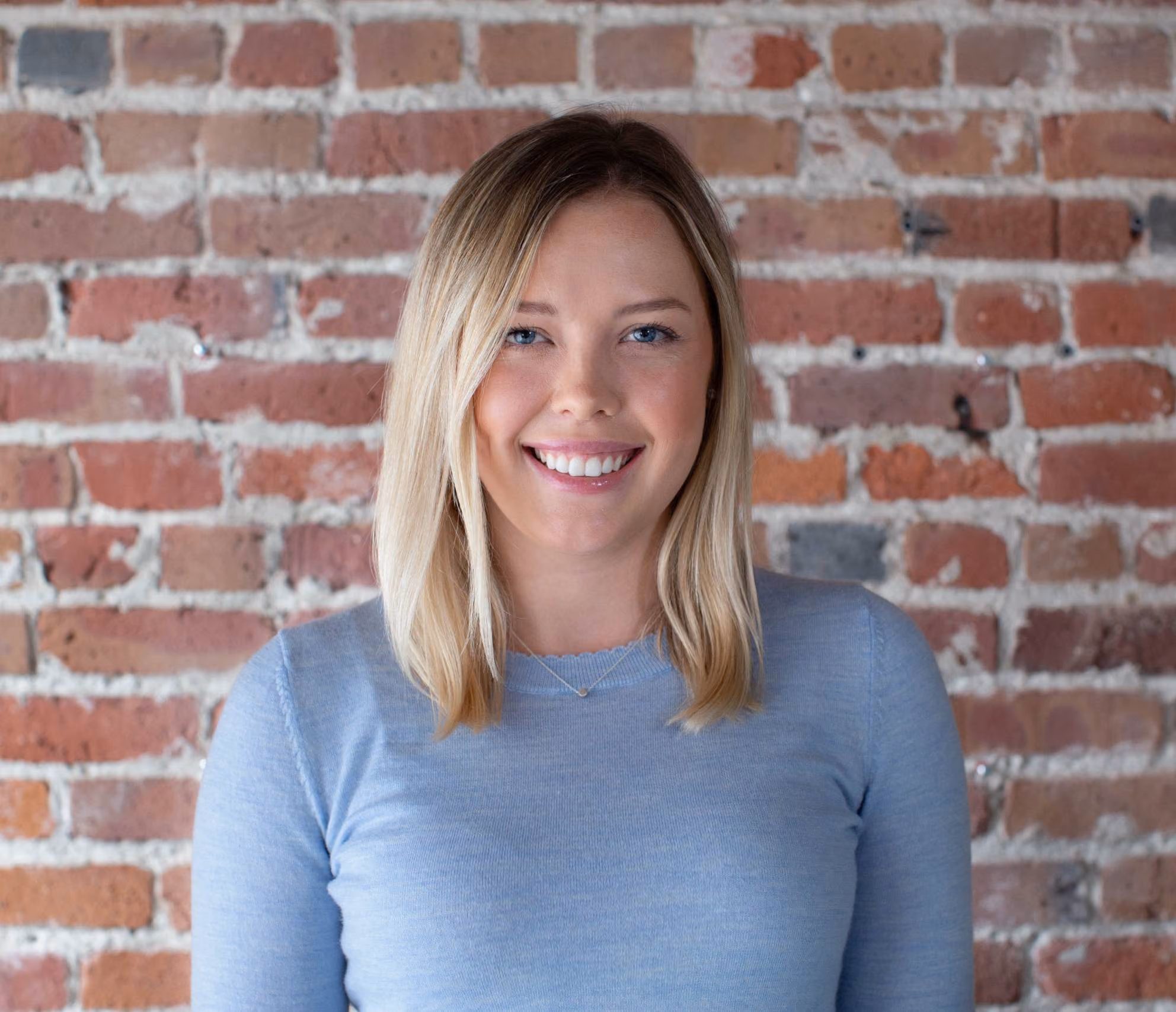 Smiling young woman with blonde hair in a light blue sweater standing in front of a brick wall.
