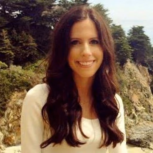 Smiling woman with long dark hair wearing a white top, outdoors with trees and rocks behind her.