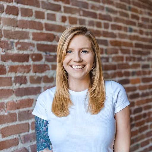 Smiling woman with blonde hair and tattoo on arm standing against a brick wall.