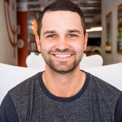 Smiling man with short dark hair and beard wearing a dark gray t-shirt sitting indoors.