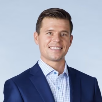 Smiling man with short hair wearing a navy blazer and checkered shirt against a light gray background.