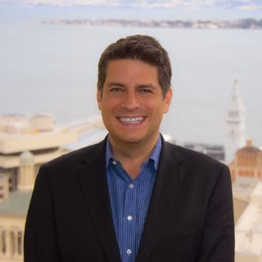 Smiling man with dark hair in blue shirt and black blazer stands outdoors with waterfront cityscape in background.