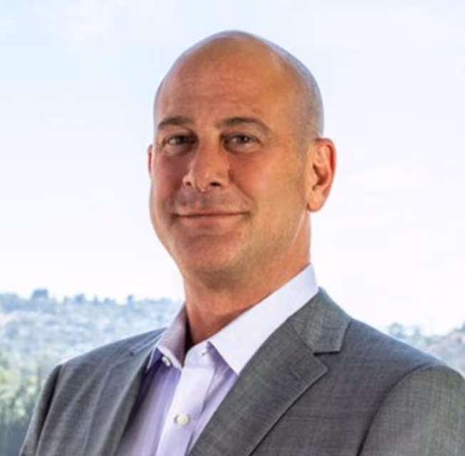 Bald man in a gray suit jacket and light purple shirt standing in front of a window with a city view.