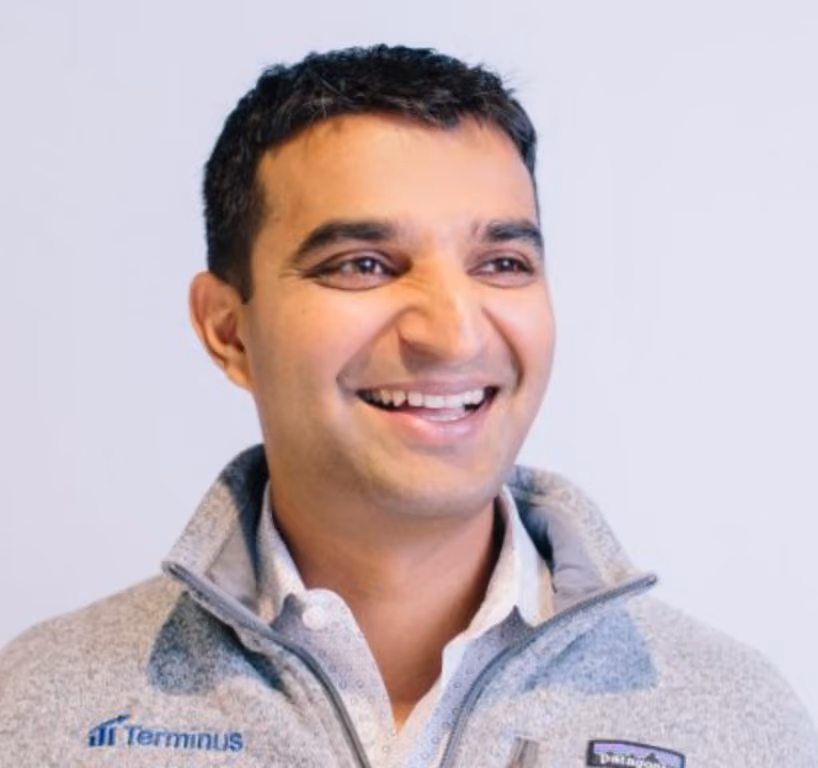 Smiling man with short dark hair wearing a gray Terminus fleece jacket over a collared shirt.