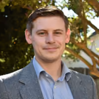Smiling man with short brown hair in a gray blazer and blue shirt outdoors with trees in the background.