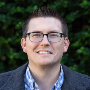 Smiling man with short hair and glasses wearing a blue checkered shirt and gray jacket outdoors.