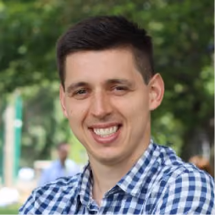 Smiling young man with short dark hair wearing a blue and white checkered shirt outdoors.