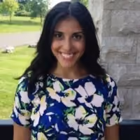 Smiling woman with long dark hair in a floral dress standing outdoors near a stone wall.
