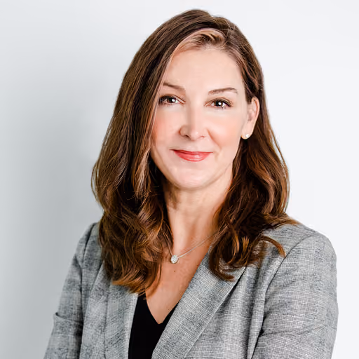 Smiling woman with shoulder-length brown hair wearing a gray blazer and black top against a white background.