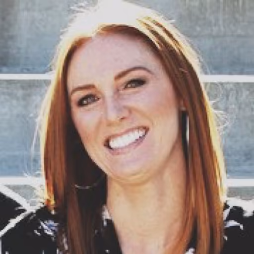 Smiling woman with long red hair and hoop earrings sitting outdoors on concrete steps.